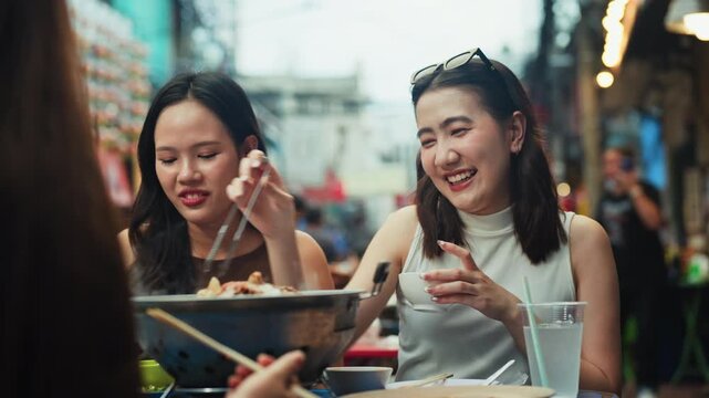 Two asian women laughing and enjoying delicious street food together at an outdoor market, sharing a fun moment with joyful expressions and vibrant atmosphere of local culture