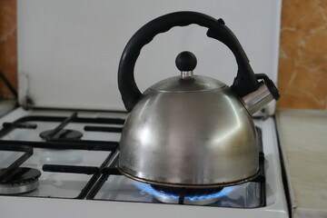 Shiny Stainless Steel Whistling Kettle Heating Water on a Lit Gas Burner with a Blue Flame on a White Kitchen Stove