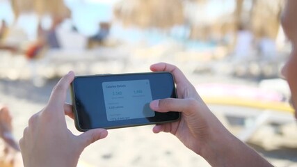 A man relaxes on the beach while using his phone to monitor his fitness progress, specifically analyzing data related to calories burned during his activities. This footage is great for sports apps