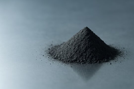 Black graphite powder heap on a smooth gray surface, lit softly to highlight texture and matte details