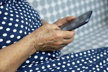 Elderly woman in polka dot dress sitting with smartphone indoors, mobile phone in wrinkled female hands closeup. Concept of online communication in retirement