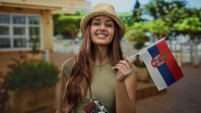 Woman smiling with camera and serbian flag in hand on an outdoor street wearing summer hat representing travel and tourism vibes in a cultural setting during daytime.