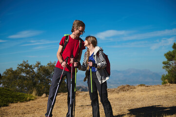 A joyful couple outdoors on a clear day, sharing a smile while hiking with walking poles. Backpacks...