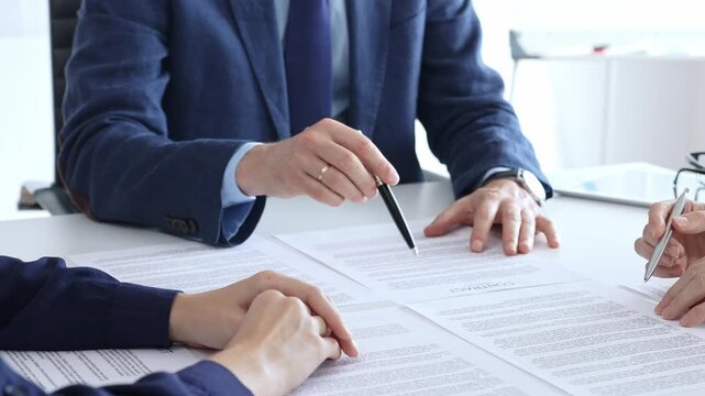 Business professionals gather in a bright office around a desk strewn with legal documents and pens, reviewing paperwork and preparing to sign a formal contract after negotiations