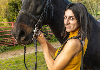 Woman smiling while adjusting a black horse's bridle. Lifestyle. Horse riding.