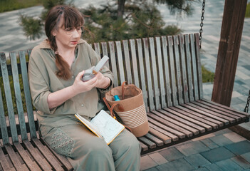 Elderly woman puts on headphones, draws in notebook, pursues hobby, sitting on bench in city park. Active age, interesting life and creative abilities of older people