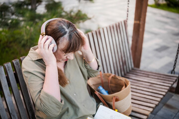 Elderly woman puts on headphones, draws in notebook, pursues hobby, sitting on bench in city park. Active age, interesting life and creative abilities of older people