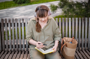 Elderly woman in headphones draws in notebook, pursues hobby, sitting on bench in city park. Active age, interesting life and creative abilities of older people