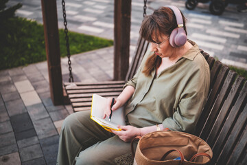 Elderly woman in headphones draws in notebook, pursues hobby, sitting on bench in city park. Active age, interesting life and creative abilities of older people
