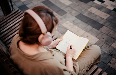 Elderly woman in headphones draws in notebook, pursues hobby, sitting on bench in city park. Active age, interesting life and creative abilities of older people