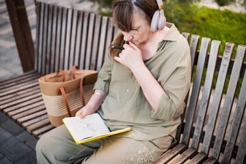 Aged woman in headphones sits thoughtfully drawing in notebook, pursues hobby, in city park. Active age, interesting life and creative abilities of older people