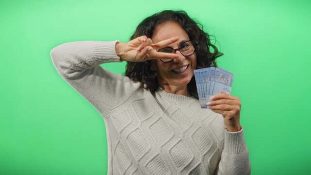 Middle-aged woman with malaysian ringgit banknotes posing playfully against a bright green wall background, making a peace sign with one hand and holding cash in the other.