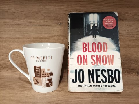A coffee cup with an Italian phrase beside a Jo Nesbo thriller novel standing on a wooden surface