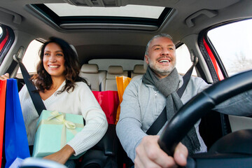 Happy man and woman driving in car with shopper bags and gifts, preparing for winter holidays on the Christmas eve