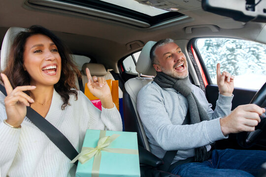 Excited spouses driving car with Xmas presents, woman holding wrapped gift box, man and woman enjoy music, singing song feeling enjoyment of winter holiday preparation