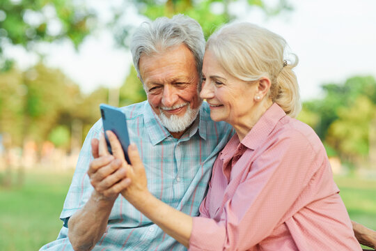 Happy active senior couple taking selfie sitting ob a bench in park outdoors. Love togetherness and acive seniors vitality concept