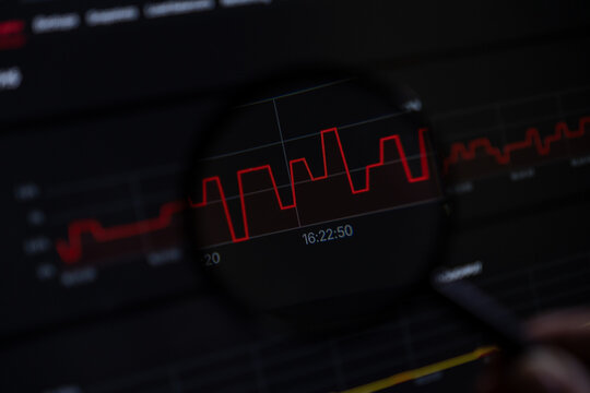Magnifying glass analyzing a volatile red line graph on screen, signifying critical data examination for market research and business intelligence insights