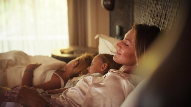 A serene morning unfolds as a mother and her children relax in a comfortable bed at a beautiful luxury resort, enjoying their family vacation.