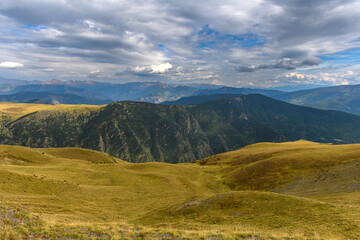 Obraz premium Foreground of a grassy mountain meadow at the end of summer, with the Spanish Pyrenees mountain range in the background. In the mid-ground, a mountain with slopes covered in pines and trees, and a sky
