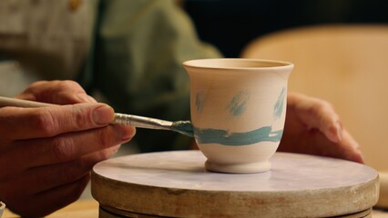Close up unknown Caucasian man male hands small business owner potter entrepreneur businessman painting ceramic clay vase pot drawing paintbrush pattern ornament design turning table indoors workshop
