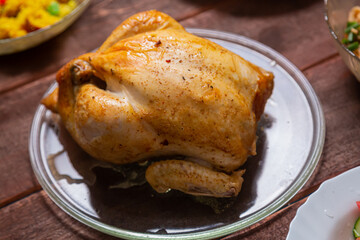Grilled whole chicken on a glass plate on a wooden table with other food. Horizontal photo.