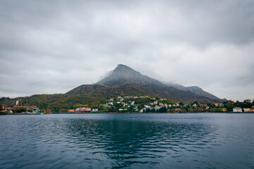 Late-afternoon view of Lake Como from Lecco, Italy, during autumn. Vibrant red, orange and green trees line the calm lakeshore beneath dramatic grey clouds. A distant couple stands by the water as the