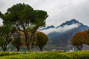 Late-afternoon view of Lake Como from Lecco, Italy, during autumn. Vibrant red, orange and green trees line the calm lakeshore beneath dramatic grey clouds. A distant couple stands by the water as the