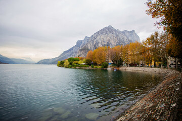 Late-afternoon view of Lake Como from Lecco, Italy, during autumn. Vibrant red, orange and green trees line the calm lakeshore beneath dramatic grey clouds. A distant couple stands by the water as the