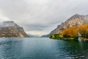 Late-afternoon view of Lake Como from Lecco, Italy, during autumn. Vibrant red, orange and green trees line the calm lakeshore beneath dramatic grey clouds. A distant couple stands by the water as the