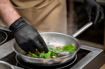 Chef cooking leafy greens and mushrooms in a steaming pan on electric stove, wearing gloves and apron in professional kitchen setting, showcasing culinary technique and hygiene.