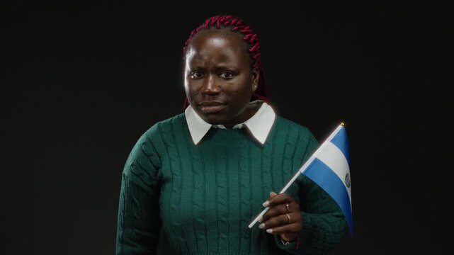 African woman holding el salvador flag with a curious expression posing against an isolated black background.