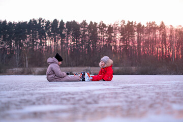 Girls in warm winter overalls relax on the surface of a frozen lake. Ice skating