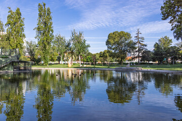 A tranquil view of the central lake in Parque do Bonfim, Set&uacute;bal, Portugal, featuring lush greenery, reflections, and ducks resting on the far bank under a bright blue sky.