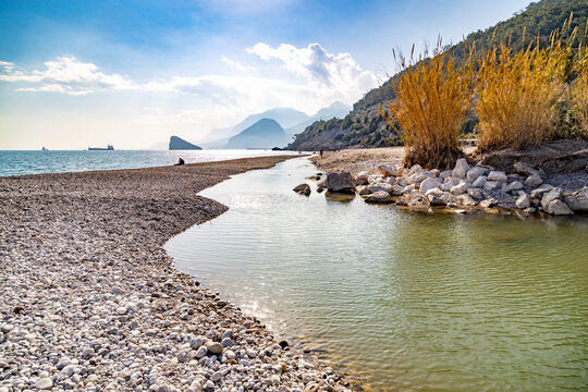 River mouth at Sarısu Beach with backlit turquoise water, silhouetted figures on shore, and yellow reeds against hazy Taurus Mountains. Winter Mediterranean landscape. Antalya, Turkey.

