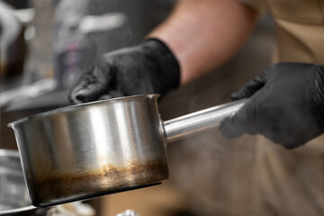 Chef holding hot stainless steel saucepan with steam rising, wearing black gloves in professional kitchen. Close-up shows signs of frequent use and heat.