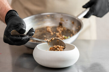 Chef plating cooked grain dish into white bowl on stainless steel surface. Hands in black gloves, apron visible. Professional kitchen, food preparation, culinary concept.