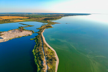 Aerial view of a contrasting landscape where a green algal bay meets a quarry lake, separated by a sandy isthmus. Surrounded by agricultural fields with yellow rapeseed, wind turbines, and sand mining