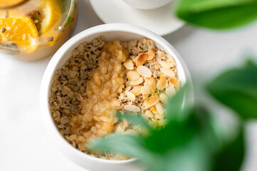 Healthy breakfast with oatmeal topped with applesauce and almonds in a white bowl. Fresh fruit jar, cup, and green plant in soft focus background. Natural light, cozy setting.
