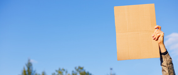 Close-up of a hand raised up and holding a blank piece of cardboard for writing against a sky background. Protest or warning. The blank space, can be used to add text or a symbol.