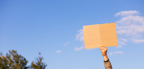 Close-up of a hand raised up and holding a blank piece of cardboard for writing against a sky background. Protest or warning. The blank space, can be used to add text or a symbol.