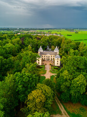 Aerial view of historic manor with twin towers, nestled in dense forest and open fields linked by a clear path and framed by distant village under dramatic overcast skies.