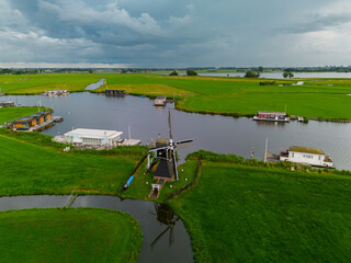Aerial view of Dutch countryside with central windmill, houseboats, canal, and green fields capturing iconic rural charm and waterway culture under dramatic overcast skies.