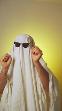 Halloween. Ghosting. Ghost costume with white blanket and sunglasses dancing over yellow background
