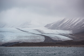A glacier with dark moraine lines descends toward the sea under low clouds on Svalbard, Norway,...