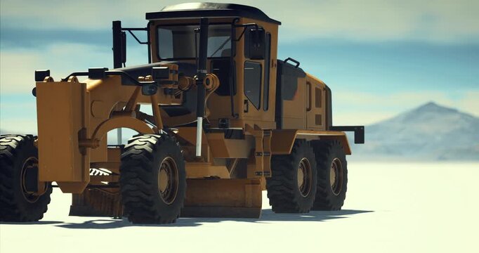 A large yellow grader navigates the white terrain, showcasing its power and precision. Snow capped mountains rise in the background, highlighting a stunning winter day in an isolated area.