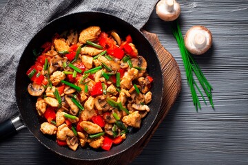 Sliced chicken and vibrant vegetables are stir-fried in a black skillet. The dish features red bell peppers and mushrooms, garnished with green onions. Fresh ingredients are ready to serve