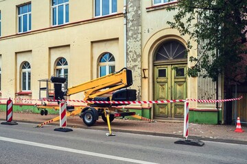Construction site with a yellow mobile lift near an old building under renovation. The area is closed with red and white safety tape, indicating ongoing repair work and facade restoration.
