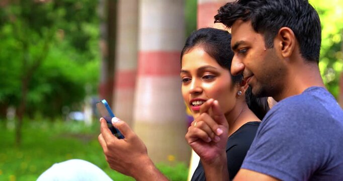 Indian beautiful young couple checking fitness app on smartphone sitting on bench in park outdoor during sunny day, tracking health progress, wellness goals, and sharing workout motivation