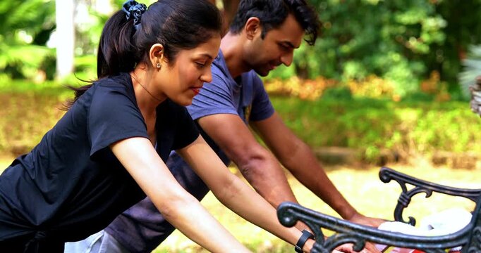 Indian beautiful young couple doing pushups on bench in park outdoors during morning fitness session, showing strength, teamwork, and healthy lifestyle while enjoying sunlight, fresh air, greenery