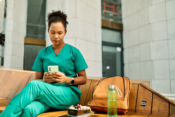 Young woman or a nurse or a doctor during office hours break enjoying a fresh salad and using mobile phone  in a park during a sunny day while smiling and relaxing on a bench